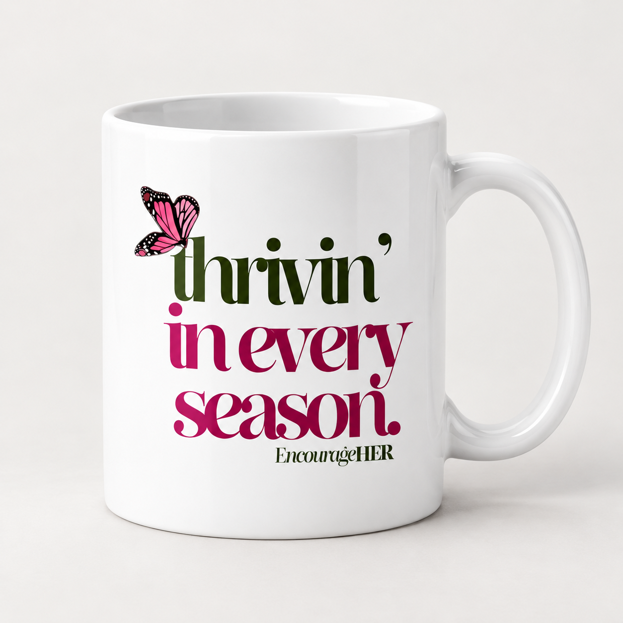 White mug with green and pink Thrivin’ in every season lettering, a pink butterfly, and EncourageHER branding